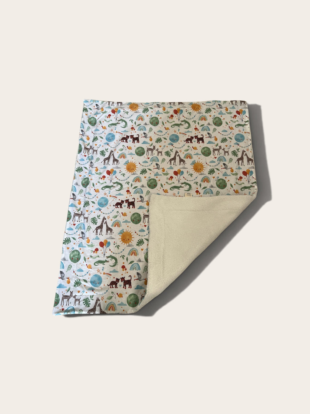 A folded blanket with a whimsical safari-themed print on one side and a soft cream fleece backing on the other, displayed on a light beige background.