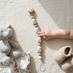 A baby pacifier clip with wooden and pastel silicone beads is held by a baby's hand on a beige linen surface, styled with a gray plush elephant toy and beige knit booties.