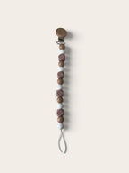 A pacifier clip with a wooden spoon-style clasp, a vertical string of brown and mauve beads, and a white loop at the bottom, shown on a solid light beige background.