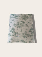 A baby blanket featuring a classic toile de Jouy pattern with palm trees, giraffes, and elephants in muted green on a warm beige linen background, shown on a solid light beige background.
