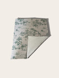 A blanket featuring a green toile de Jouy pattern with palm trees, giraffes, and elephants on a light beige background. The underside is solid light beige fleece.