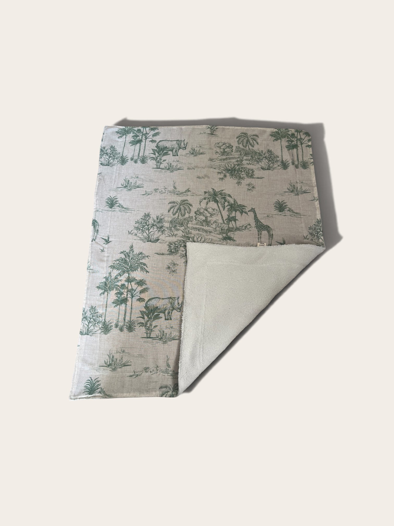 A blanket featuring a green toile de Jouy pattern with palm trees, giraffes, and elephants on a light beige background. The underside is solid light beige fleece.
