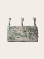 A cot organiser with a green safari toile pattern on a light beige linen-like fabric, featuring three adjustable tie straps at the top, shown against a solid off-white background.