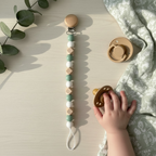 A wooden pacifier clip with green and white beads hangs on a light wood surface next to a pacifier and a baby's hand reaching for it. A green patterned blanket and eucalyptus leaves are also visible.