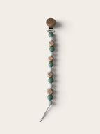 A wooden pacifier clip with a round brown wooden top, a silver metal clasp, and a dangling string of faceted wooden beads in sage green, light beige, and medium brown