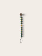 A beaded pacifier clip with sage green beads, small gold spacer beads, a white daisy accent, and a brown snap closure, shown on a solid light beige background.