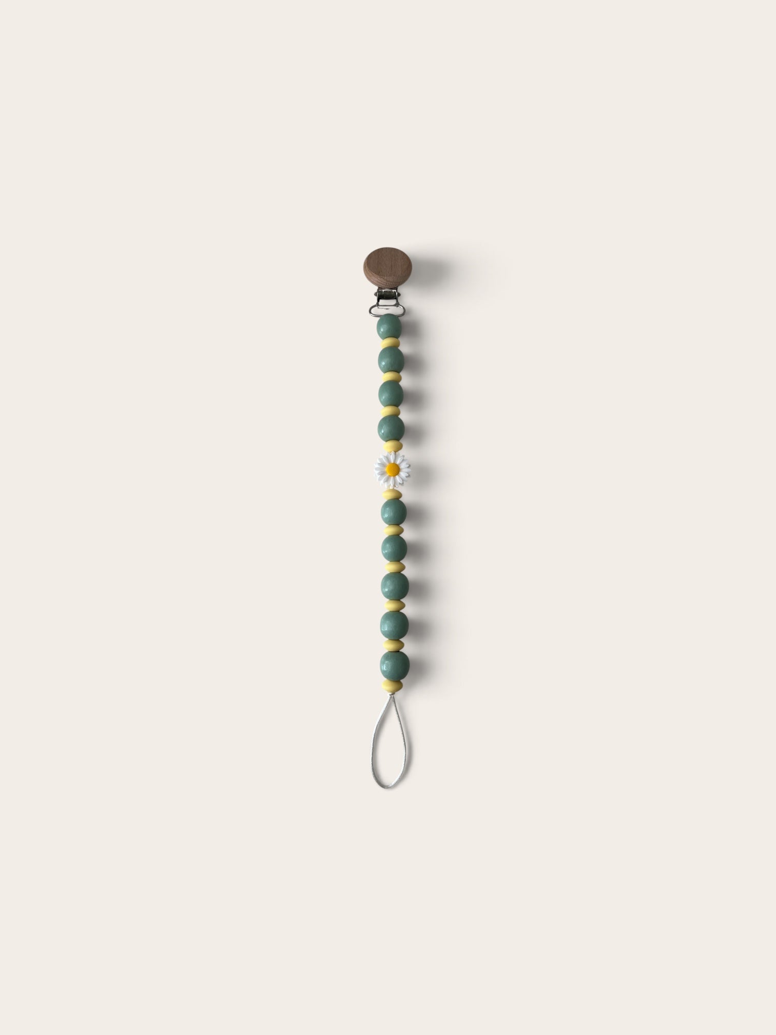 A beaded pacifier clip with sage green beads, small gold spacer beads, a white daisy accent, and a brown snap closure, shown on a solid light beige background.