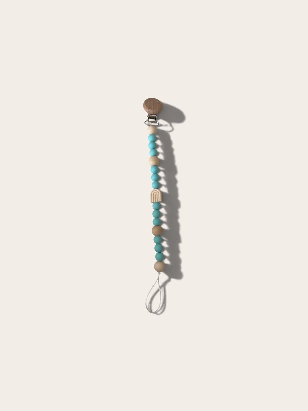 A wooden pacifier clip with turquoise and beige beads on a white cord, shown on a light beige background.