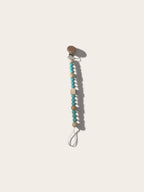 A wooden pacifier clip with turquoise and beige beads on a white cord, shown on a light beige background.