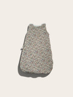 A sleeveless baby sleeping bag or pouch with a dense small floral print on a light beige background, featuring a white zipper at the top and a side zipper, shown against a solid off-white background.