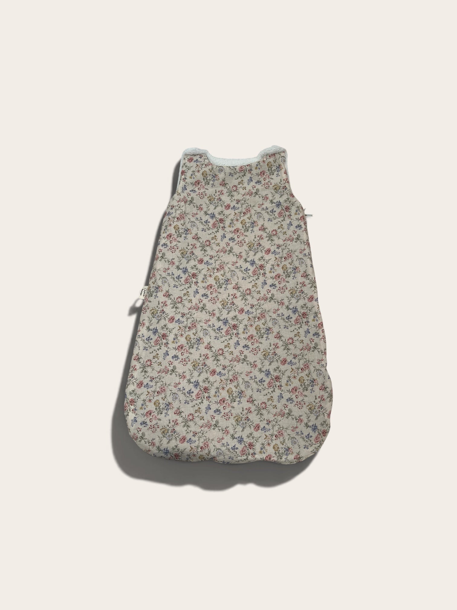 A sleeveless baby sleeping bag or pouch with a dense small floral print on a light beige background, featuring a white zipper at the top and a side zipper, shown against a solid off-white background.