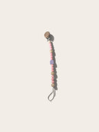 A wooden pacifier clip with pastel pink and beige beads, a small lavender striped accent bead, and a natural wood teething tab on a light cream background.